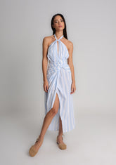 Modelo veste o Vestido Adriana em algodão 100% com listras azul, amarração no pescoço e cintura, detalhe drapeado e fenda frontal, ideal para dias quentes com frescor e sofisticação.
