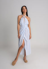 Modelo veste o Vestido Adriana em algodão 100% com listras azul, amarração no pescoço e cintura, detalhe drapeado e fenda frontal, ideal para dias quentes com frescor e sofisticação.